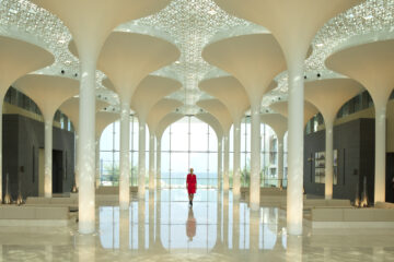 a woman in a red dress in a large room with white columns with Sheikh Zayed Mosque in the background