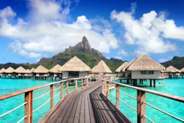 a wooden walkway leading to a beach with huts on stilts with Bora Bora in the background