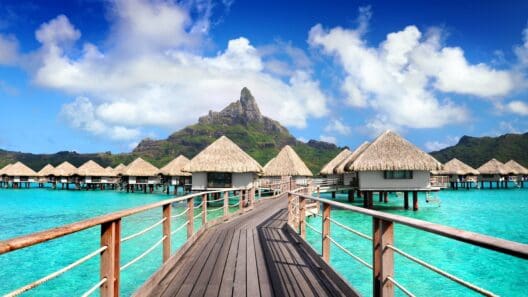 a wooden walkway leading to a beach with huts on stilts with Bora Bora in the background