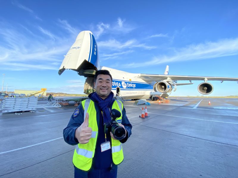 a man in a yellow vest holding a camera and standing in front of an airplane