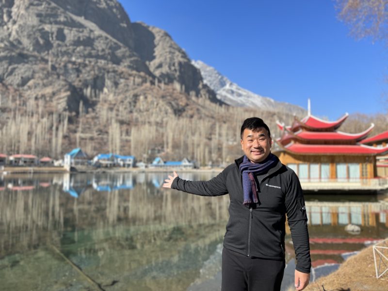 a man standing in front of a body of water with a red pagoda and mountains in the background
