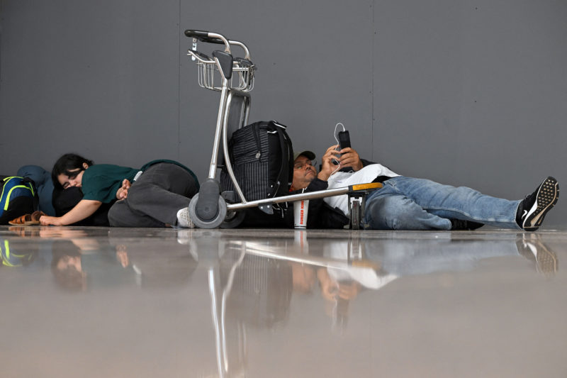 a man lying on the ground with a luggage cart and a man looking at his phone