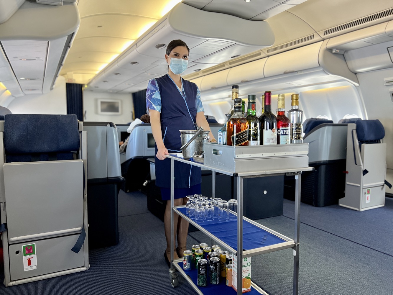 a woman wearing a face mask standing in an airplane with a cart full of bottles