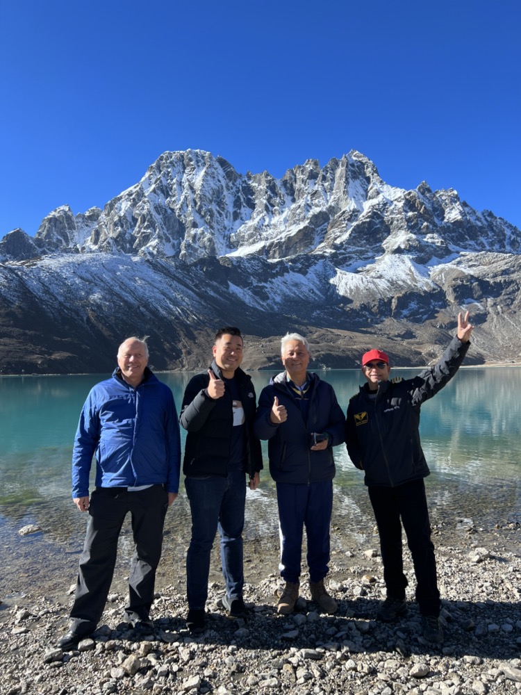a group of men standing in front of a lake with snow covered mountains in the background