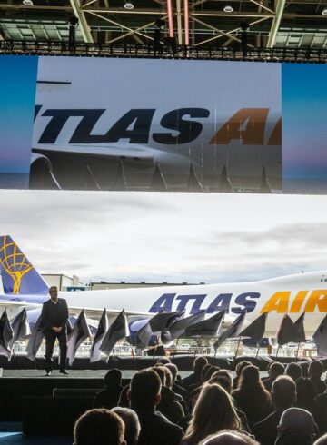 a man on a stage with a crowd watching a large screen with airplanes