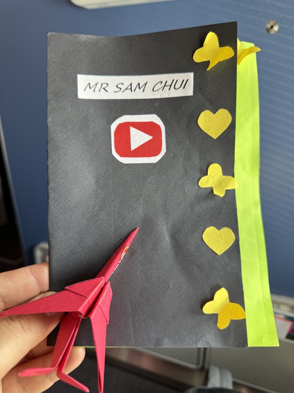 a hand holding a grey envelope with a red paper airplane and yellow paper flowers