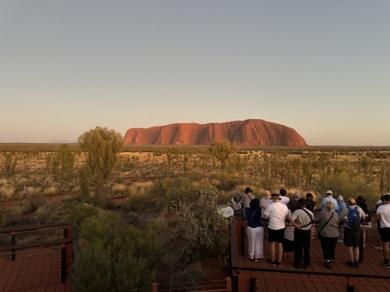 a group of people standing on a deck looking at a large rock formation with Uluru in the background