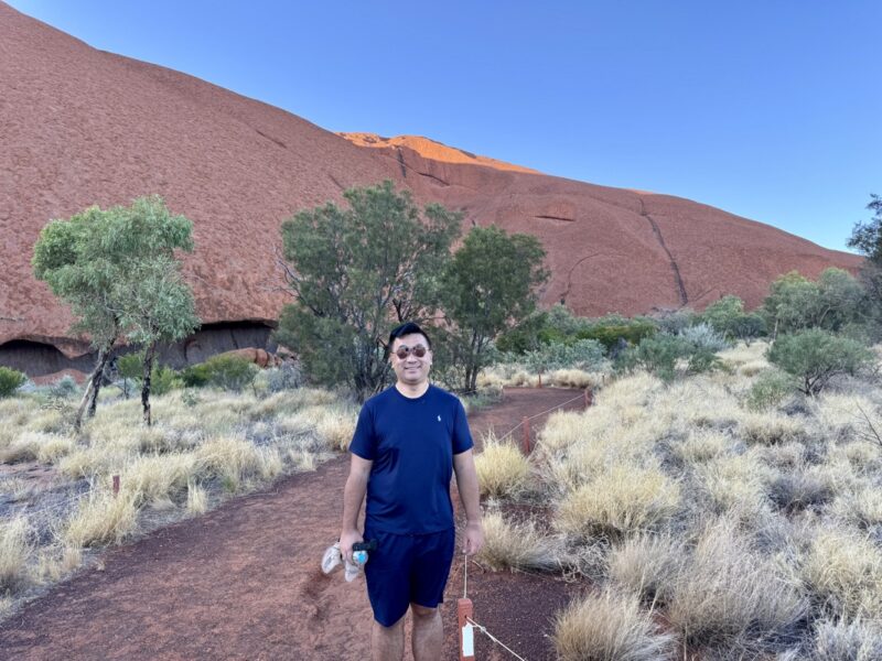 a man standing in a dirt path with a large red rock in the background