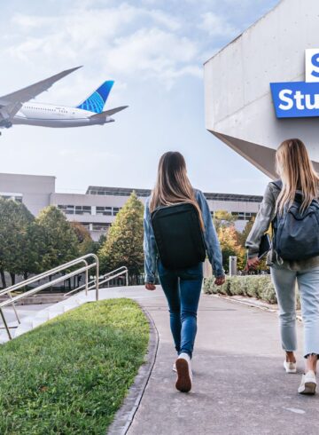 two women walking on a sidewalk with a plane flying in the sky